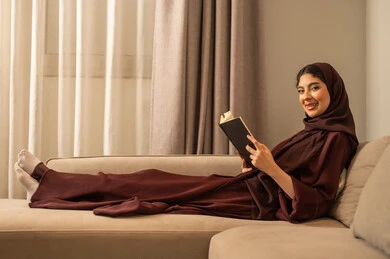قضاء الوقت بالقراءة والتعلم، ثقافة القراءة والمعرفة، امراة عربية خليجية سعودية محجبة ترتدي العباءة تجلس على الاريكة  و تقوم بقراءة الكتاب، الجلوس في غرفة المعيشة