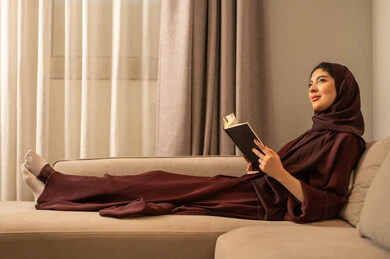 The culture of reading and knowledge, a Saudi Arabian Gulf woman in a hijab wearing an abaya sits on the sofa and reads a book, spending time reading and learning, sitting in the living room.