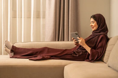 Completing work tasks via a tablet, the technical application for the tablet, online shopping, accomplishing tasks remotely, using modern and advanced devices and technologies, an Arab Gulf Saudi woman wearing a hijab and an abaya sitting on the sofa in the living room.