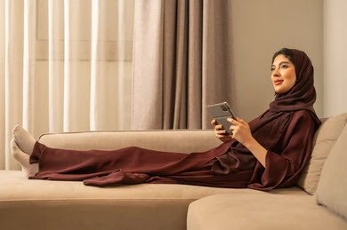 التطبيق التقني  للجهاز اللوحي، التسوق الالكتروني عبر الأونلاين، إنجاز المهام عن بعد، استخدام الأجهزة والتقنيات الحديثة والمتطورة, إمرأة عربية خليجية سعودية محجبة ترتدي العباءة  تجلس على الاريكة فى غرفه المعيشة تقوم بإنجاز مهام العمل عبر جهاز التابلت