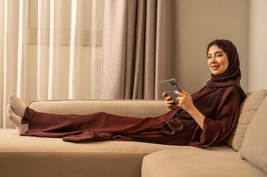 التسوق الالكتروني عبر الأونلاين، إنجاز المهام عن بعد، التطبيقات التقنية للأجهزة اللوحية، استخدام الأجهزة والتقنيات الحديثة والمتطورة, إمرأة عربية خليجية سعودية محجبة ترتدي العباءة  تجلس على الاريكة فى غرفه المعيشة تقوم بإنجاز مهام العمل عبر جهاز التابلت