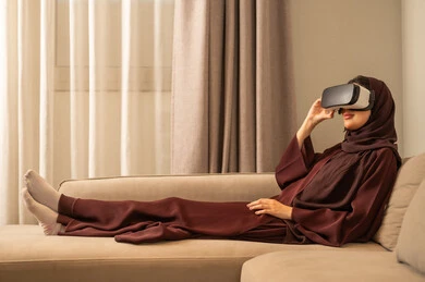 Integration and interaction with the virtual world, using modern electronic technology and devices, enjoying leisure time, an Arab Gulf Saudi woman wearing a hijab sitting on the couch wearing 3D VR glasses.
