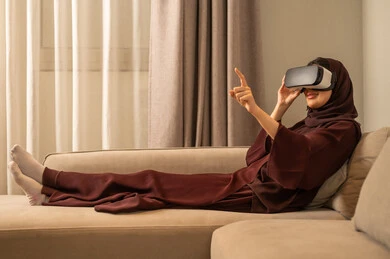 الاشارة الى شيء ما، استخدام تقنية حديثة الكترونية وأجهزة تكنولوجية، الاستمتاع في قضاء الوقت الترفيهي، امراة عربية خليجية سعودية محجبة تجلس على الاريكه تضع نظارات الفي آر ثلاثية الابعاد، الاندماج والتفاعل مع العالم الافتراضي