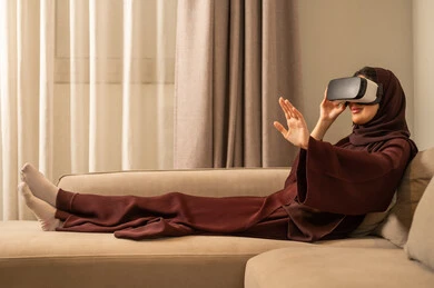 استخدام تقنية حديثة الكترونية وأجهزة تكنولوجية، الاستمتاع في قضاء الوقت الترفيهي، امراة عربية خليجية سعودية محجبة تجلس على الاريكه تضع نظارات الفي آر ثلاثية الابعاد، الاندماج والتفاعل مع العالم الافتراضي، الاشارة الى شيء ما