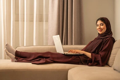Professionalism in computer and information technology jobs, remote work and browsing the internet, using modern and advanced technical devices, a photo of an Arab Gulf Saudi woman wearing an abaya and hijab sitting on the sofa at home, using a laptop, completing work tasks.