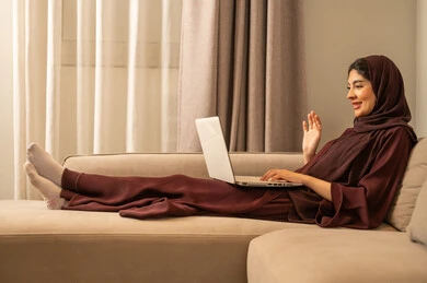Raising the hand in a gesture of peace and greeting, making a video call, working remotely and browsing the internet, using modern and advanced technological devices, a photo of an Arab Gulf Saudi woman wearing an abaya and hijab sitting on the sofa at home, using a laptop, completing work tasks.