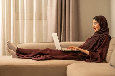 العمل عن بعد وتصفح الانترنت، احتراف وظائف الحاسوب وتكنولوجيا المعلومات، استخدام الأجهزة التقنية الحديثة و المتطورة ،تصوير لامرأة عربية خليجية سعودية ترتدي العباءة والحجاب تجلس على الاريكة في المنزل ، استخدام جهاز الحاسوب المحمول، انجاز مهام العمل