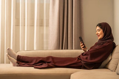 تصفح مواقع التواصل الاجتماعي عبر الجوال، امراه عربية خليجية سعودية ترتدي الحجاب والعباءة تنظر تجلس على الأريكة في غرفة المعيشة و تمسك بيديها الهاتف المحمول ، تعابير السعادة و السرور