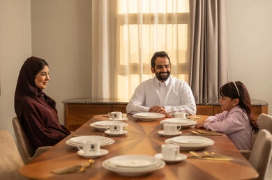 Weekend, having a good time, expressions of happiness and joy, a Saudi Arabian Gulf couple wearing traditional attire sitting at the dining table at home with their daughter, a happy Arab Gulf family, a joyful family atmosphere.