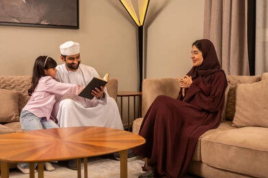 Enhancing the family bond, the concept of fatherhood and childhood, spending enjoyable time with children, an Arab Gulf Omani man wearing a dishdasha and kumma sitting in the living room with his daughter and wife reading entertaining stories, developing the hobby of reading.