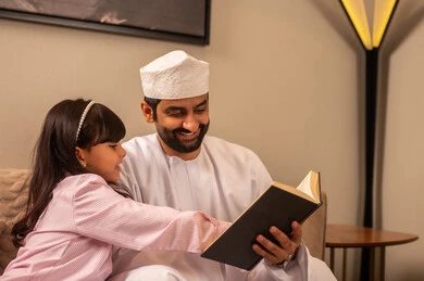 The concept of fatherhood and sonship, spending quality time with children, an Arab Gulf Omani man wearing a dishdasha and kumma sitting in the living room with his daughter reading entertaining stories, strengthening the family bond, developing the hobby of reading.