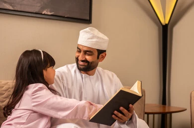 قضاء وقت ممتع مع الأبناء ، مفهوم الابوة والبنوة، رجل عربي خليجي عماني يرتدي الدشداشة و الكمة يجلس فى غرفه المعيشة مع ابنته يقوم بقراءة القصص المسلية، تعزيز الروابط الاسرية، تنمية هواية المطالعة