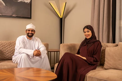 The concept of family bonding and understanding, affection and love between spouses, an Arab Gulf Omani man wearing a dishdasha and a kumma sitting in the living room with his wife, looking at the camera with expressions of happiness and joy.