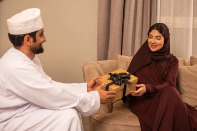 Gestures of happiness and joy, expressing intimacy and affection between spouses, an Arab Gulf Omani man wearing a dishdasha and kumma presents a gift to his wife on the occasion of Valentine's Day, celebrating occasions and holidays.