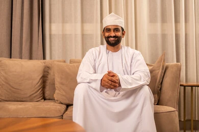 النظر الى الكاميرا بتعابير السعادة و السرور، الأناقة والاهتمام بالمظهر الخارجي،  الجلوس على الاريكة ايدي متشابكة ، مفهوم الرجولة والثقة بالنفس, رجل عربي خليجي عماني  يرتدي  الدشداشة و الكمة يجلس فى غرفه المعيشة