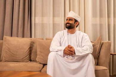 الأناقة والاهتمام بالمظهر الخارجي،  الجلوس على الاريكة ايدي متشابكة ، مفهوم الرجولة والثقة بالنفس, رجل عربي خليجي عماني  يرتدي  الدشداشة و الكمة يجلس فى غرفه المعيشة ، النظر الى شيء ما