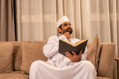 قضاء الوقت بالقراءة والتعلم والثقافة، تعابير التفكير فى شيء ما، ايماءات التمعن والتركيز، مفهوم التعلم الذاتي، رجل عربي خليجي عماني يرتدي الدشداشة و الكمة يجلس على الاريكة فى غرفه المعيشة ويحمل بيده كتاب