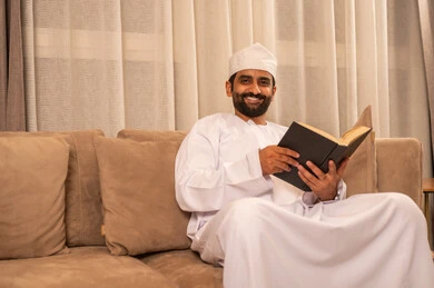 الاهتمام بتصفح الكتب المفيدة،  قضاء وفت الفراغ بالتثقيف والتعلم، تنمية مهارة القراءة، رجل عربي خليجي عماني  يرتدي الدشداشة و الكمة يجلس على الاريكة فى غرفه المعيشة ويحمل بيده كتاب