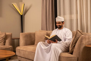 Spending free time on education and learning, developing reading skills, paying attention to browsing useful books, an Arab Gulf Omani man wearing a dishdasha and a kumma sits on the sofa in the living room holding a book in his hand.