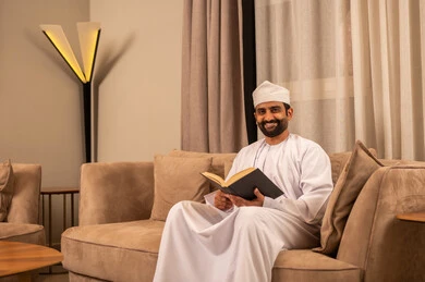 تنمية مهارة القراءة، قضاء وفت الفراغ بالتثقيف والتعلم, الاهتمام بتصفح الكتب المفيدة، رجل عربي خليجي عماني  يرتدي الدشداشة و الكمة يجلس على الاريكة فى غرفه المعيشة ويحمل بيده كتاب