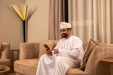Browsing social media, chatting with family and friends, using a modern technological device, an Arab Gulf Omani man wearing a dishdasha and kumma sits on the sofa in the living room holding a mobile phone in his hand, merging technology and technology into daily life.