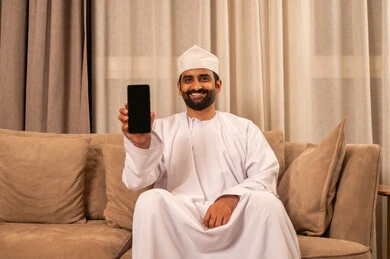 Using a modern technical device, displaying a black screen on the mobile phone, chatting with family and friends, an Arab Gulf Omani man wearing a dishdasha and kumma sitting on the sofa in the living room holding a mobile phone in his hand, integrating technology and tech into daily life, browsing social media, expressions of happiness and joy.