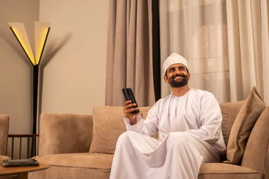 الدردشة مع الأهل والأصدقاء، استخدام جهاز تقني حديث، رجل عربي خليجي عماني يرتدي الدشداشة و الكمة يجلس على الاريكة فى غرفه المعيشة يمسك بيده الهاتف المحمول، دمج التقنية والتكنولوجيا بالحياة اليومية، تصفح مواقع التواصل الاجتماعي، النظر الى شيء ما