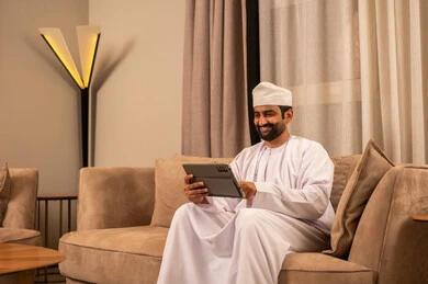 استخدام تطبيقات و برنامج الجهاز اللوحي ، الدردشة مع الأهل والأصدقاء، تعابير السعادة و السرور، استعمال التكنولوجيا والتقنيات الحديثة، رجل عربي خليجي عماني يرتدي الدشداشة و الكمة يجلس على الاريكة فى غرفه المعيشة يحمل بيده جهاز التابلت
