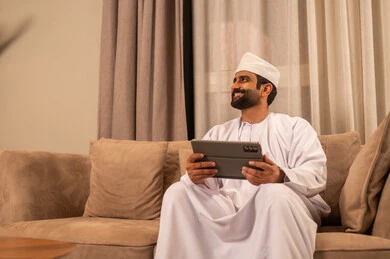 الدردشة مع الأهل والأصدقاء، النظر الى شيء ما، استعمال التكنولوجيا والتقنيات الحديثة، رجل عربي خليجي عماني يرتدي الدشداشة و الكمة يجلس على الاريكة فى غرفه المعيشة يحمل بيده جهاز التابلت، استخدام تطبيقات وبرامج الأجهزة اللوحية