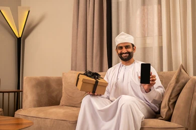 عرض شاشة سوداء عبر الهاتف المحمول، تقديم الهدايا بالمناسبات العائلية السعيدة, زيادة روابط المحبة بالإهداء والتقدير,شاب عربي خليجي عماني يرتدي الدشداشة و الكمة يجلس فى غرفة المعيشة يمسك بيده علبة هدية ملفوفة بشريط أنيق و الجوال, تعابير السعادة و السرور