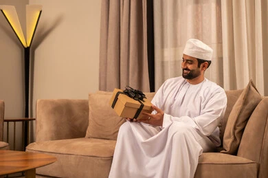 أعياد الميلاد والمباركات والأفراح، تقديم الهدايا بالمناسبات العائلية السعيدة, زيادة روابط المحبة بالإهداء والتقدير,شاب عربي خليجي عماني يرتدي الدشداشة و الكمة يجلس فى غرفة المعيشة يمسك بيده علبة هدية ملفوفة بشريط أنيق, تعابير السعادة و السرور