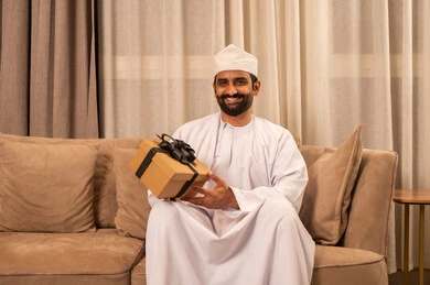 تقديم الهدايا بالمناسبات العائلية السعيدة, أعياد الميلاد والمباركات والأفراح, زيادة روابط المحبة بالإهداء والتقدير,شاب عربي خليجي عماني يرتدي الدشداشة و الكمة يجلس فى غرفة المعيشة يمسك بيده علبة هدية ملفوفة بشريط أنيق, تعابير السعادة و السرور