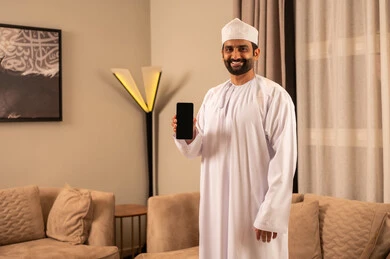 عرض شاشة سوداء فارغه عبر الجوال، رجل عربي خليجي عماني يرتدي الدشداشة و الكمة يقف فى غرفه المعيشة  يمسك بيدية الهاتف المحمول ، تعابير السعادة و السرور