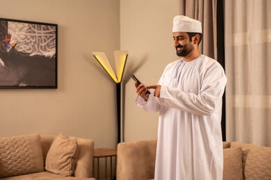 تصفح موقع التواصل الاجتماعي، رجل عربي خليجي عماني يرتدي الدشداشة و الكمة يقف فى غرفه المعيشة  يمسك بيدية الهاتف المحمول ، تعابير السعادة و السرور