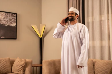 The use of modern technology, an Arab Gulf Omani man wearing a dishdasha and kumma stands in the living room making a phone call via his mobile phone, with expressions of happiness and joy. The use of modern technology, an Arab Gulf Omani man wearing a dishdasha and kumma stands in the living room making a phone call via his mobile phone, with expressions of happiness and joy.
