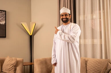 Pointing to something with gestures of joy, expressions of assurance and determination, an Arab Gulf Omani man wearing a dishdasha and a kumma stands in the living room looking at the camera, with expressions of happiness and joy.