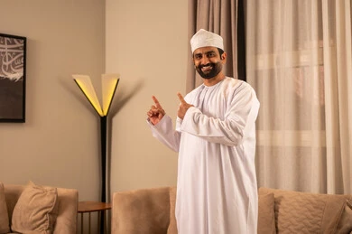 Expressions of certainty and specification, an Arab Gulf Omani man wearing a dishdasha and kumma stands in the living room looking at the camera, pointing to something with gestures of joy, expressions of happiness and delight.