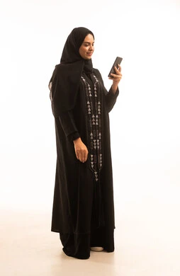 استخدام جهاز تقني حديث ومتطور، النظر الى شىء ما، بورتريه لامرأة عربية خليجية سعودية محجبة ترتدي العباءة السوداء تمسك بيدها الهاتف المحمول، الجسم بالطول الكامل، خلفية بيضاء