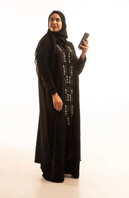 Looking at something, a portrait of a Saudi Arabian Gulf woman wearing a black abaya and a hijab, holding a mobile phone in her hand, using a modern and advanced technological device, full-body, white background.