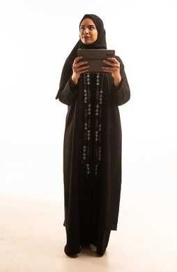Accomplishing tasks remotely, online shopping, using modern and advanced devices and technologies, a portrait of a Saudi Arabian Gulf woman wearing a black abaya and a hijab using a tablet, full-body, white background.