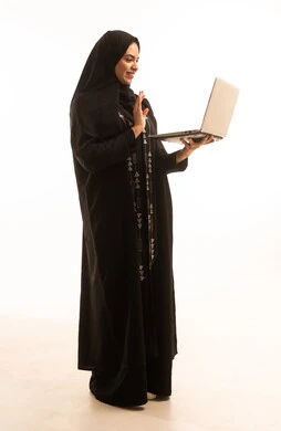 دمج التكنولوجيا في الحياة العملية، الدردشة والتواصل مع الاهل والاصدقاء عبر الجهاز التقني الحديث، العمل عن بعد وتصفح الانترنت باستخدام الكومبيوتر،بورتريه لامرأة عربية خليجية سعودية محجبة ترتدي عباءة سوداء تمسك بيدها الحاسوب، خلفية بيضاء