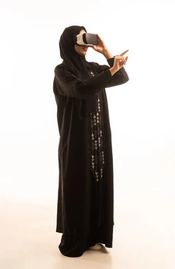 المتعة والتسلية بالمشاهدة عبر نظارة الواقع الافتراضي، بورتريه من الجانب لإمرأة عربية خليجية سعودية محجبة ترتدي العباءة السوداء تضع نظارات ثلاثية الأبعاد تشير باصبع السبابة الى شىء ما, ايماءات السعادة والسرور، خلفية بيضاء