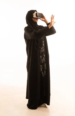 A side portrait of a Saudi Arabian Gulf woman wearing a black abaya and a hijab, wearing 3D glasses and pointing with her index finger at something. The scene depicts immersion and simulation of a fantasy world, enjoyment and entertainment through watching via virtual reality glasses, with gestures of happiness and joy, against a white background.