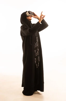 الاندماج ومحاكاة العالم الخيالي، المتعة والتسلية بالمشاهدة عبر نظارة الواقع الافتراضي, بورتريه من الجانب لإمرأة عربية خليجية سعودية محجبة ترتدي العباءة السوداء تضع نظارات ثلاثية الأبعاد تشير باصبع السبابة الى شىء ما, ايماءات السعادة والسرور، خلفية بيضاء