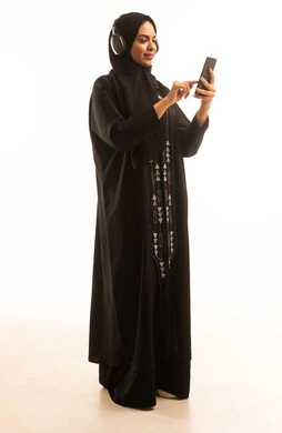 بورتريه لامرأة عربية خليجية سعودية محجبة ترتدى عباءة سوداء تضع الهيدفون تحمل بيدها الهاتف المحمول، عرض شاشة سوداء فارغة عبر الجوال،استخدام جهاز تقني حديث ومتطور، الجسم بالطول الكامل، خلفية بيضاء