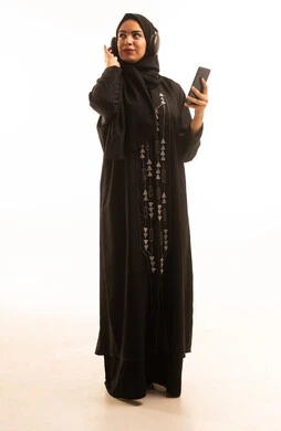 Using a modern and advanced technical device, a portrait of a Saudi Arabian Gulf woman wearing a black hijab and an abaya, wearing headphones and holding a mobile phone in her hand, displaying a blank black screen on the phone, looking at the camera with gestures of joy, full-body shot, white background. Using a modern and advanced technical device, a portrait of a Saudi Arabian Gulf woman wearing a black hijab and an abaya, wearing headphones and holding a mobile phone in her hand, displaying a blank black screen on the phone, looking at the camera with gestures of joy, full-body shot, white background.