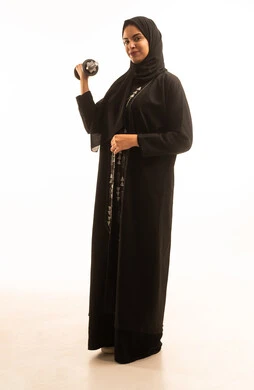 A portrait of a smiling Saudi Arabian Gulf woman wearing a black abaya and a hijab, holding heavy iron weights (dumbbells) in her hand, looking at the camera with gestures of confidence, embodying the concept of fitness, flexibility, and physical agility, following a healthy lifestyle, engaging in sports activities, with a white background.