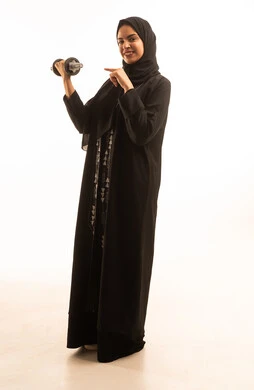 مفهوم اللياقة والليونة والرشاقة البدنية, اتباع أسلوب حياة صحي, ممارسة الأنشطة الرياضية, بورتريه لسيدة عربية خليجية سعودية محجبة مبتسمة ترتدي العباءة السوداء تحمل بيدها الاوزان الثقيلة الحديدية ( الدمبلز) تنظر إلى الكاميرا بإيماءات الثقة, خلفية بيضاء