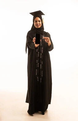عرض شاشة سوداء فارغة، إيماءات السعادة بالوصول لحلم النجاح، بورتريه لخريجة عربية خليجية سعودية محجبة ترتدي عباءة سوداء والقبعة تمسك بيدها هاتف محمول، الاحتفال بإنهاء الدراسة الجامعية، الجسم بالطول الكامل، خلفية بيضاء
