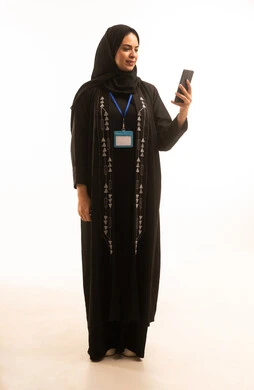 Using a smartphone application, a portrait of a smiling Saudi Arabian Gulf woman wearing a black abaya and holding a white card with a blue ribbon while holding a mobile phone, with a white background. Using a smartphone application, a portrait of a smiling Saudi Arabian Gulf woman wearing a black abaya and holding a white card with a blue ribbon while holding a mobile phone, with a white background.